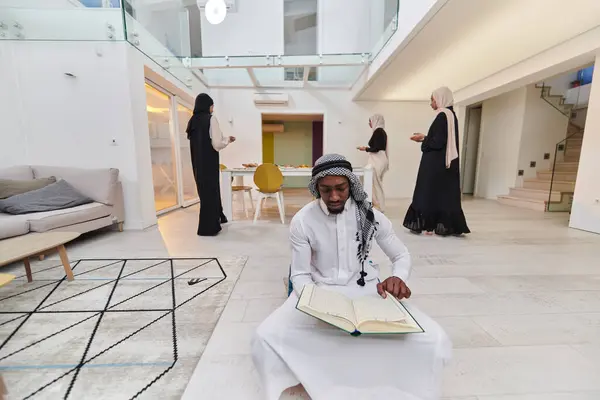 In the sacred month of Ramadan, an African American Muslim man engrossed in reading the Holy Quran is surrounded by a backdrop where three hijab-wearing women prepare and serve food for iftar