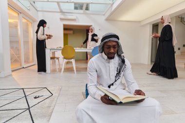 Kutsal Ramazan ayında, Kuran okumaya dalmış Afrikalı bir Müslüman adam üç tesettürlü kadının iftar için yemek hazırlayıp servis ettiği bir zemin ile çevrilidir.