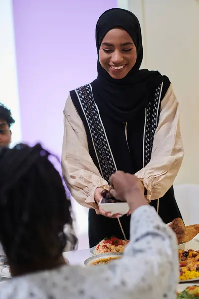 In a heartwarming scene during the sacred month of Ramadan, a traditional Muslim woman offers dates to her family gathered around the table, exemplifying the spirit of unity, generosity, and cultural