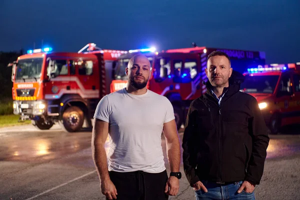 Group of firefighters, dressed in civilian clothing, stand in front of fire trucks during the night, showcasing a moment of camaraderie and unity among the team as they reflect on their duties and the