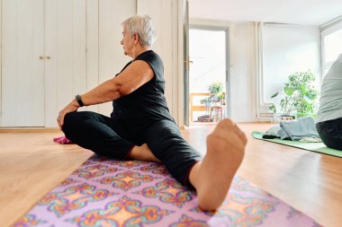 Yaşlı bir kadın çeşitli yoga pozlarına katılıyor, uzuvlarını esnetiyor ve eğitimli bir eğitmenin rehberliğinde modern bir güneş ışığında huzur buluyor.