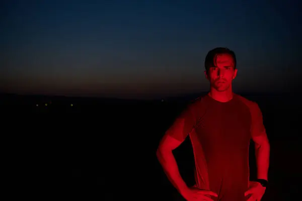 stock image In the solemn darkness illuminated by a red glow, an athlete strikes a confident pose, embodying resilience and determination after completing a grueling day-long marathon.