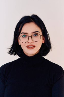 Esmer bir kadın, gözlüğünü gururla takarken neşe saçıyor. Temiz, beyaz bir arka plana karşı sıcak bir gülümseme sergiliyor.