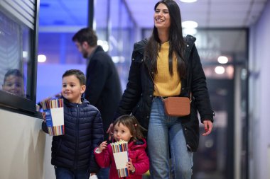 A young couple with their children stands outside the cinema, purchasing freshly popped popcorn before the start of the movie and entry into the theater. clipart