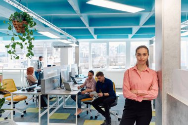 Gencecik gülümseyen iş kadınının portresi. Yaratıcı açık mekan ortak çalışma ofisinde. Başarı sahibi iş kadını, kopyalama alanı ile ofiste duruyor. Arka planda çalışan iş arkadaşları. 