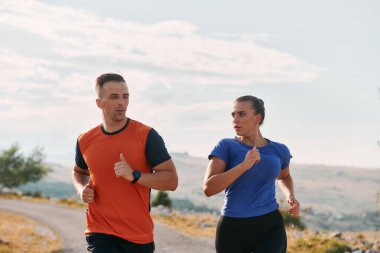 Spor kıyafetleri giymiş bir çift sabahın erken saatlerinde spor yaparken manzaralı bir yolda koşuyor, temiz havanın tadını çıkarıyor ve sağlıklı bir yaşam tarzı sürdürüyorlar..