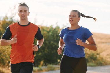 Spor kıyafetleri giymiş bir çift sabahın erken saatlerinde spor yaparken manzaralı bir yolda koşuyor, temiz havanın tadını çıkarıyor ve sağlıklı bir yaşam tarzı sürdürüyorlar..