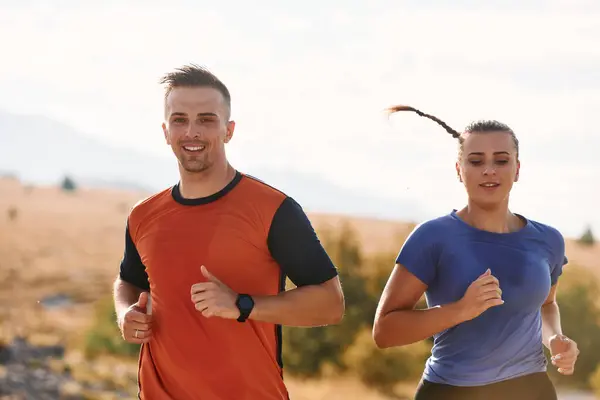 stock image A couple dressed in sportswear runs along a scenic road during an early morning workout, enjoying the fresh air and maintaining a healthy lifestyle.
