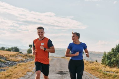 Spor kıyafetleri giymiş bir çift sabahın erken saatlerinde spor yaparken manzaralı bir yolda koşuyor, temiz havanın tadını çıkarıyor ve sağlıklı bir yaşam tarzı sürdürüyorlar..