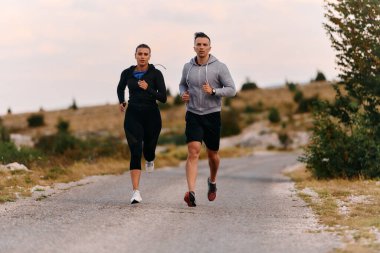 A couple dressed in sportswear runs along a scenic road during an early morning workout, enjoying the fresh air and maintaining a healthy lifestyle. clipart