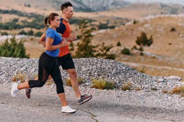 Spor kıyafetleri giymiş bir çift sabahın erken saatlerinde spor yaparken manzaralı bir yolda koşuyor, temiz havanın tadını çıkarıyor ve sağlıklı bir yaşam tarzı sürdürüyorlar..