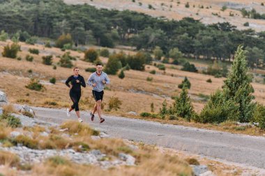 Spor kıyafetleri giymiş bir çift sabahın erken saatlerinde spor yaparken manzaralı bir yolda koşuyor, temiz havanın tadını çıkarıyor ve sağlıklı bir yaşam tarzı sürdürüyorlar..