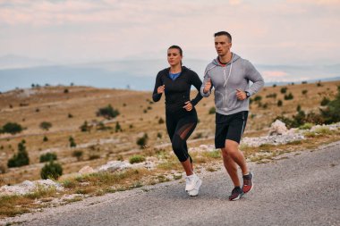 Spor kıyafetleri giymiş bir çift sabahın erken saatlerinde spor yaparken manzaralı bir yolda koşuyor, temiz havanın tadını çıkarıyor ve sağlıklı bir yaşam tarzı sürdürüyorlar..
