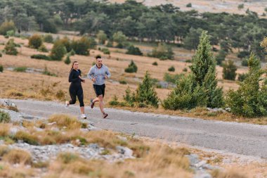Spor kıyafetleri giymiş bir çift sabahın erken saatlerinde spor yaparken manzaralı bir yolda koşuyor, temiz havanın tadını çıkarıyor ve sağlıklı bir yaşam tarzı sürdürüyorlar..