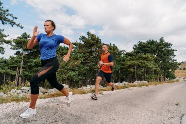 Spor kıyafetleri giymiş bir çift sabahın erken saatlerinde spor yaparken manzaralı bir yolda koşuyor, temiz havanın tadını çıkarıyor ve sağlıklı bir yaşam tarzı sürdürüyorlar..