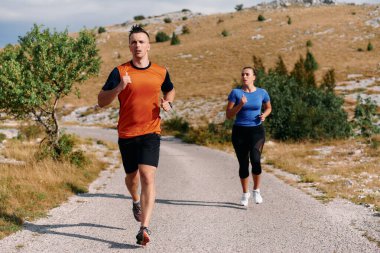 Spor kıyafetleri giymiş bir çift sabahın erken saatlerinde spor yaparken manzaralı bir yolda koşuyor, temiz havanın tadını çıkarıyor ve sağlıklı bir yaşam tarzı sürdürüyorlar..