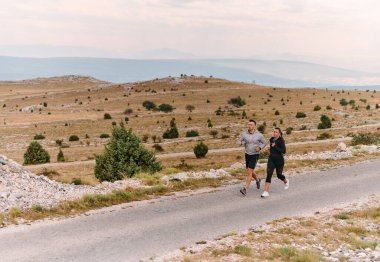 Spor kıyafetleri giymiş bir çift sabahın erken saatlerinde spor yaparken manzaralı bir yolda koşuyor, temiz havanın tadını çıkarıyor ve sağlıklı bir yaşam tarzı sürdürüyorlar..