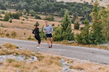 Spor kıyafetleri giymiş bir çift sabahın erken saatlerinde spor yaparken manzaralı bir yolda koşuyor, temiz havanın tadını çıkarıyor ve sağlıklı bir yaşam tarzı sürdürüyorlar..