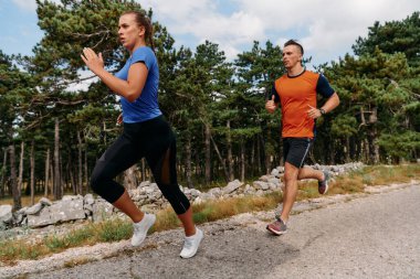 Spor kıyafetleri giymiş bir çift sabahın erken saatlerinde spor yaparken manzaralı bir yolda koşuyor, temiz havanın tadını çıkarıyor ve sağlıklı bir yaşam tarzı sürdürüyorlar..