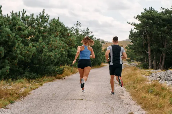 Spor kıyafetleri giymiş bir çift sabahın erken saatlerinde spor yaparken manzaralı bir yolda koşuyor, temiz havanın tadını çıkarıyor ve sağlıklı bir yaşam tarzı sürdürüyorlar..