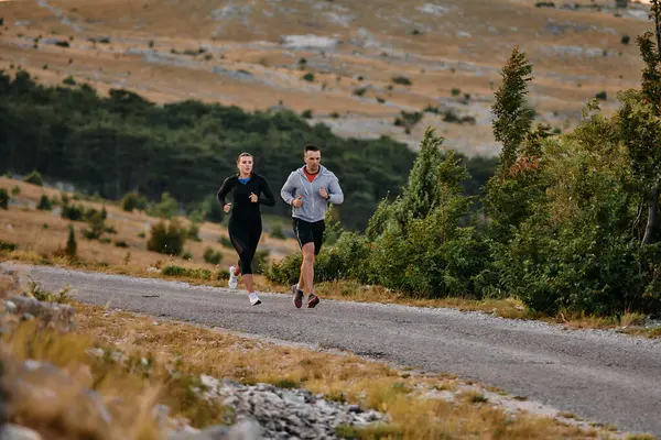 Spor kıyafetleri giymiş bir çift sabahın erken saatlerinde spor yaparken manzaralı bir yolda koşuyor, temiz havanın tadını çıkarıyor ve sağlıklı bir yaşam tarzı sürdürüyorlar..