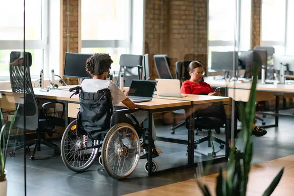 車椅子のアフリカ系アメリカ人の実業家を含む若いビジネスグループは コンピュータとラップトップの周りに積極的に従事し 集合的に解決する現代のガラスオフィス内で協力しています ストックフォト