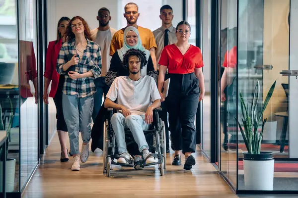 一群形形色色的年轻人聚集在一个玻璃封闭的现代创业办公室里 与坐在轮椅上的一个人 一个非洲裔美国年轻人和一个头巾都是包容性的 免版税图库图片