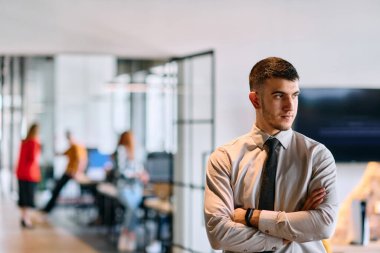 Genç bir iş adamı modern bir ofis koridorunda kollarını kavuşturarak duruyor. Kendine güven ve bir amaç duygusu yayıyor. Dinamik ve ilham verici bir varlığı somutlaştırıyor.. 