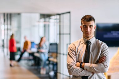 Genç bir iş adamı modern bir ofis koridorunda kollarını kavuşturarak duruyor. Kendine güven ve bir amaç duygusu yayıyor. Dinamik ve ilham verici bir varlığı somutlaştırıyor.. 