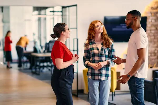 stock image A group of young business individuals, including a girl with orange hair and an African American man, stands in a modern corporate hallway, collectively examining business progress on a smartphone