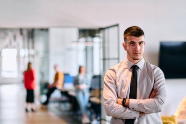 Genç bir iş adamı modern bir ofis koridorunda kollarını kavuşturarak duruyor. Kendine güven ve bir amaç duygusu yayıyor. Dinamik ve ilham verici bir varlığı somutlaştırıyor.. 