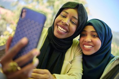 Türbanla süslenmiş iki Ortadoğulu Müslüman kadın, doğal bir ortamda otururken akıllı bir telefondan selfie çekerken bir anlık dostluk ve neşe yakalıyorlar.. 