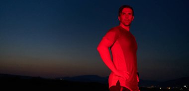 Kızıl bir parıltıyla aydınlatılan kutsal karanlıkta bir sporcu, yorucu bir gün boyunca süren maratonu tamamladıktan sonra özgüvenli bir duruş sergiler ve kararlılığını somutlaştırır..
