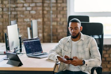  Afrikalı Amerikalı girişimci, sosyal medyayı taramak için akıllı telefon kullanarak modern bir ofise ara veriyor, iş çabaları arasında dijital bir bağlantı ve rahatlama anı yakalıyor