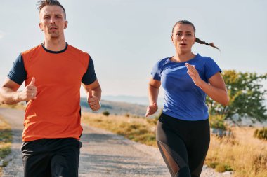 Spor kıyafetleri giymiş bir çift sabahın erken saatlerinde spor yaparken manzaralı bir yolda koşuyor, temiz havanın tadını çıkarıyor ve sağlıklı bir yaşam tarzı sürdürüyorlar..