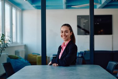 Pembe gömlekli başarılı bir bayan lider modern cam bir ofiste oturmuş kararlı bir gülümsemeyle bekliyor.