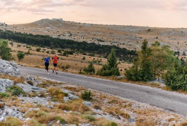 Spor kıyafetleri giymiş bir çift sabahın erken saatlerinde spor yaparken manzaralı bir yolda koşuyor, temiz havanın tadını çıkarıyor ve sağlıklı bir yaşam tarzı sürdürüyorlar..