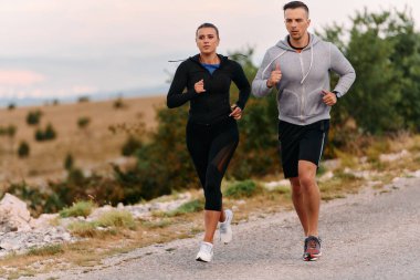 Spor kıyafetleri giymiş bir çift sabahın erken saatlerinde spor yaparken manzaralı bir yolda koşuyor, temiz havanın tadını çıkarıyor ve sağlıklı bir yaşam tarzı sürdürüyorlar..