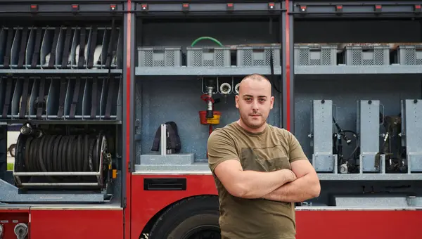 stock image Confident firefighter stands with crossed arms, exuding resilience and preparedness, ready to respond to emergencies alongside a modern fire truck, showcasing the heroism and strength of the fire