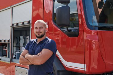 Kendine güvenen itfaiyeci, çapraz kollarla ayakta duruyor, dayanıklılık ve hazırlık yayıyor, modern bir itfaiye aracının yanında acil durumlara cevap vermeye hazır, ateşin kahramanlığını ve gücünü gösteriyor.