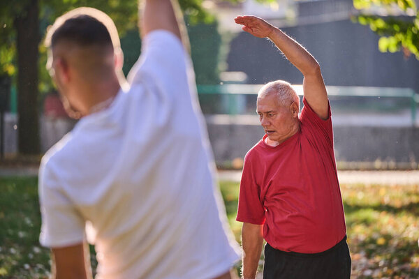 A group of seniors follows a trainer, engaging in outdoor exercises in the park, as they collectively strive to maintain vitality and well-being, embracing an active and health-conscious lifestyle in
