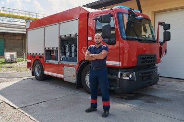 Kendine güvenen itfaiyeci, çapraz kollarla ayakta duruyor, dayanıklılık ve hazırlık yayıyor, modern bir itfaiye aracının yanında acil durumlara cevap vermeye hazır, ateşin kahramanlığını ve gücünü gösteriyor.