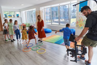 Small nursery school children with female teacher on floor indoors in classroom, doing exercise. Jumping over hula hoop circles track on the floor clipart