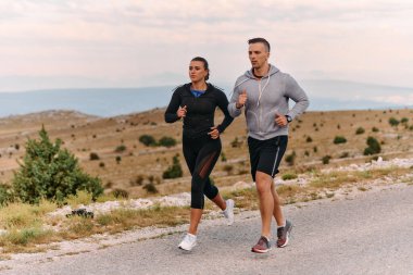 Spor kıyafetleri giymiş bir çift sabahın erken saatlerinde spor yaparken manzaralı bir yolda koşuyor, temiz havanın tadını çıkarıyor ve sağlıklı bir yaşam tarzı sürdürüyorlar..
