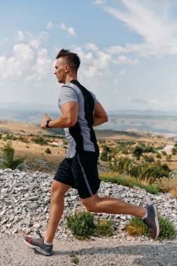Kaslı bir erkek sporcu gün doğumunda engebeli bir dağ yolunda koşar, nefes kesen kayalık manzaralar ve doğal güzelliklerle çevrilidir.. 