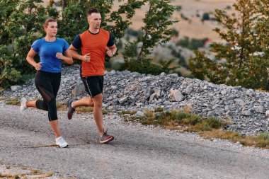 Spor kıyafetleri giymiş bir çift sabahın erken saatlerinde spor yaparken manzaralı bir yolda koşuyor, temiz havanın tadını çıkarıyor ve sağlıklı bir yaşam tarzı sürdürüyorlar..