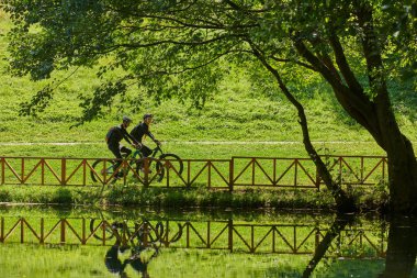 Profesyonel bisiklet kıyafetleriyle süslenmiş mutlu bir çift, parkın içinde romantik bir bisiklet sürmekten hoşlanıyor, modern doğal cazibelerle çevrili, sevgi ve mutluluk saçıyor..