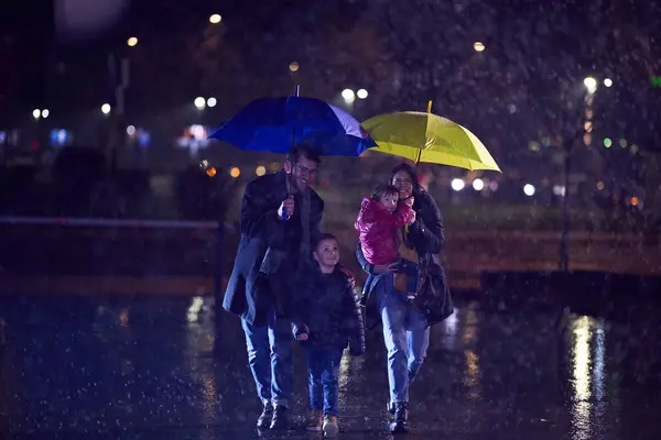 stock image In the midst of a rainy urban night, a happy couple takes their children on a stroll through the city streets, heading towards the cinema for a delightful family movie outing.