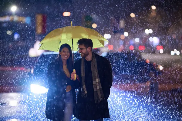 stock image In the romantic ambiance of a rainy night, a happy couple walks through the city, sharing tender moments under a yellow umbrella, surrounded by the glistening glow of urban lights.