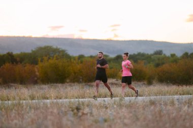 Spor kıyafetleri giymiş bir çift sabahın erken saatlerinde spor yaparken manzaralı bir yolda koşuyor, temiz havanın tadını çıkarıyor ve sağlıklı bir yaşam tarzı sürdürüyorlar.
